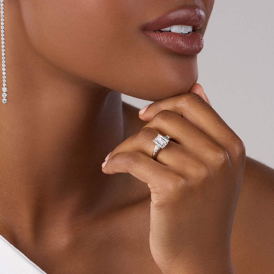 woman wearing graduated diamond dangle earrings with an emerald cut three stone engagement ring with baguettes